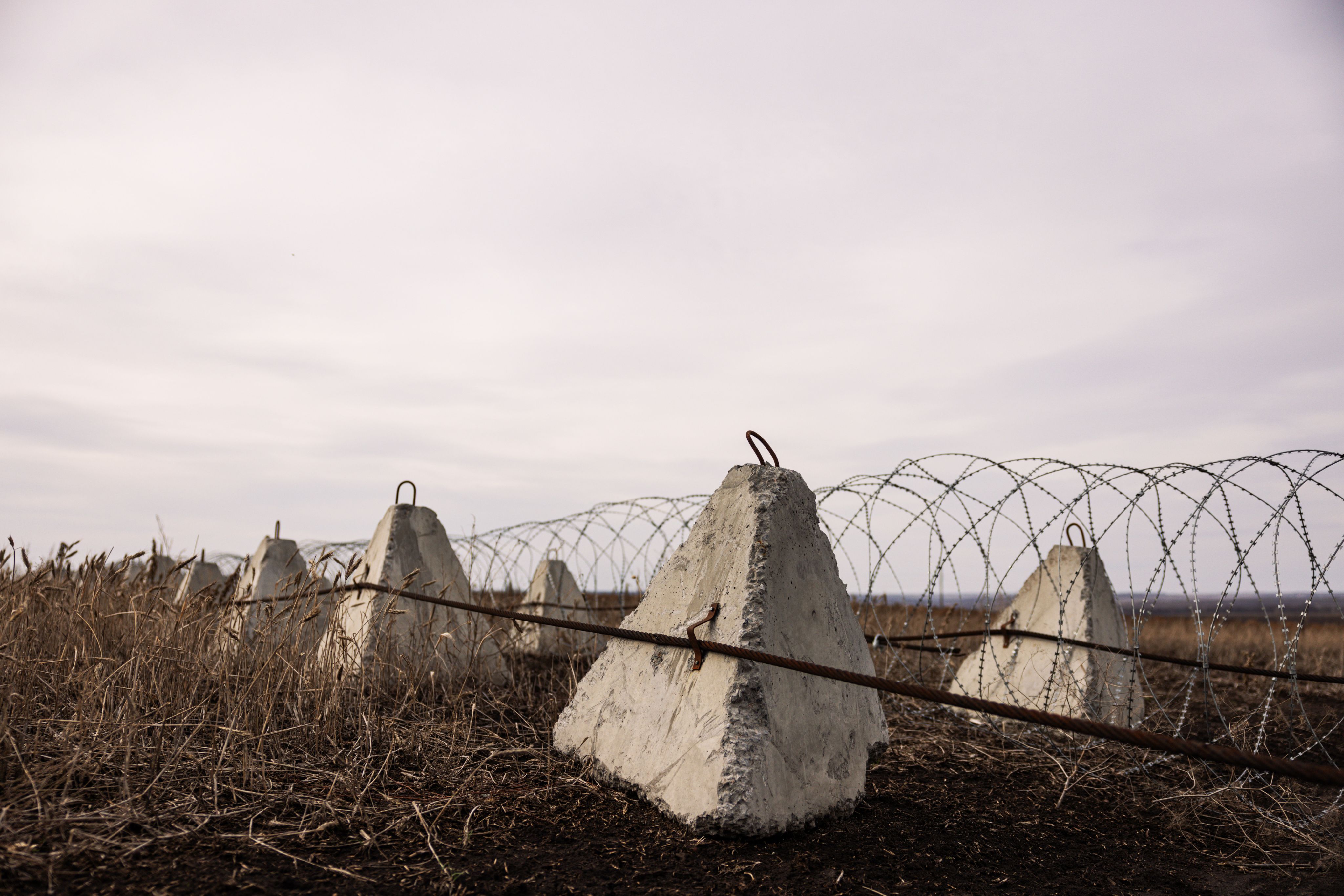 Een rij zogenoemde drakentanden, betonnen blokken bedoeld om tanks te stoppen. Ertussen zit prikkeldraad. De drakentanden en prikkeldraad liggen op Oekraïens landbouwgebied.